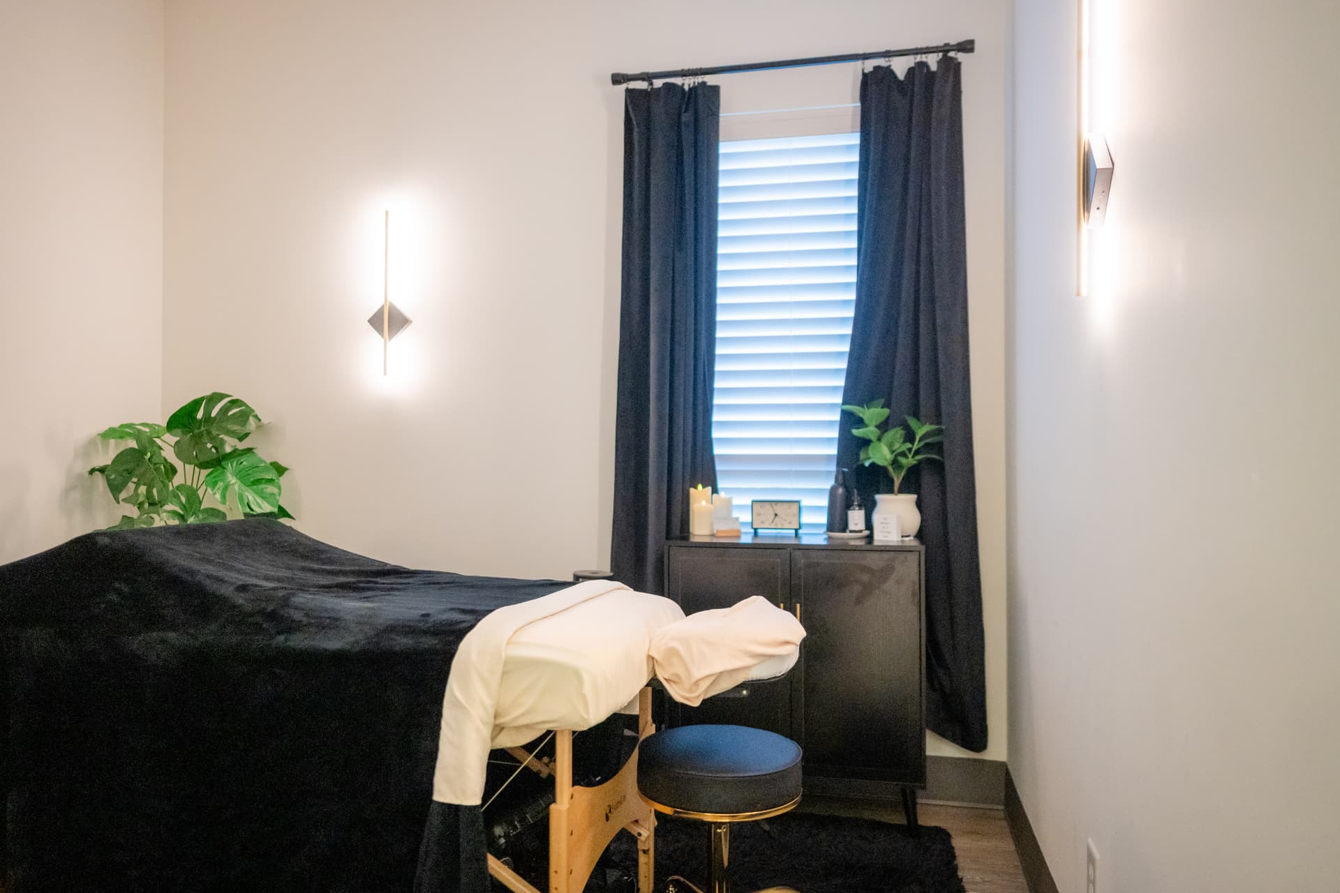 A softly lit massage treatment room at Restora with a black table, cream linens, dark curtains, plants, and candles
