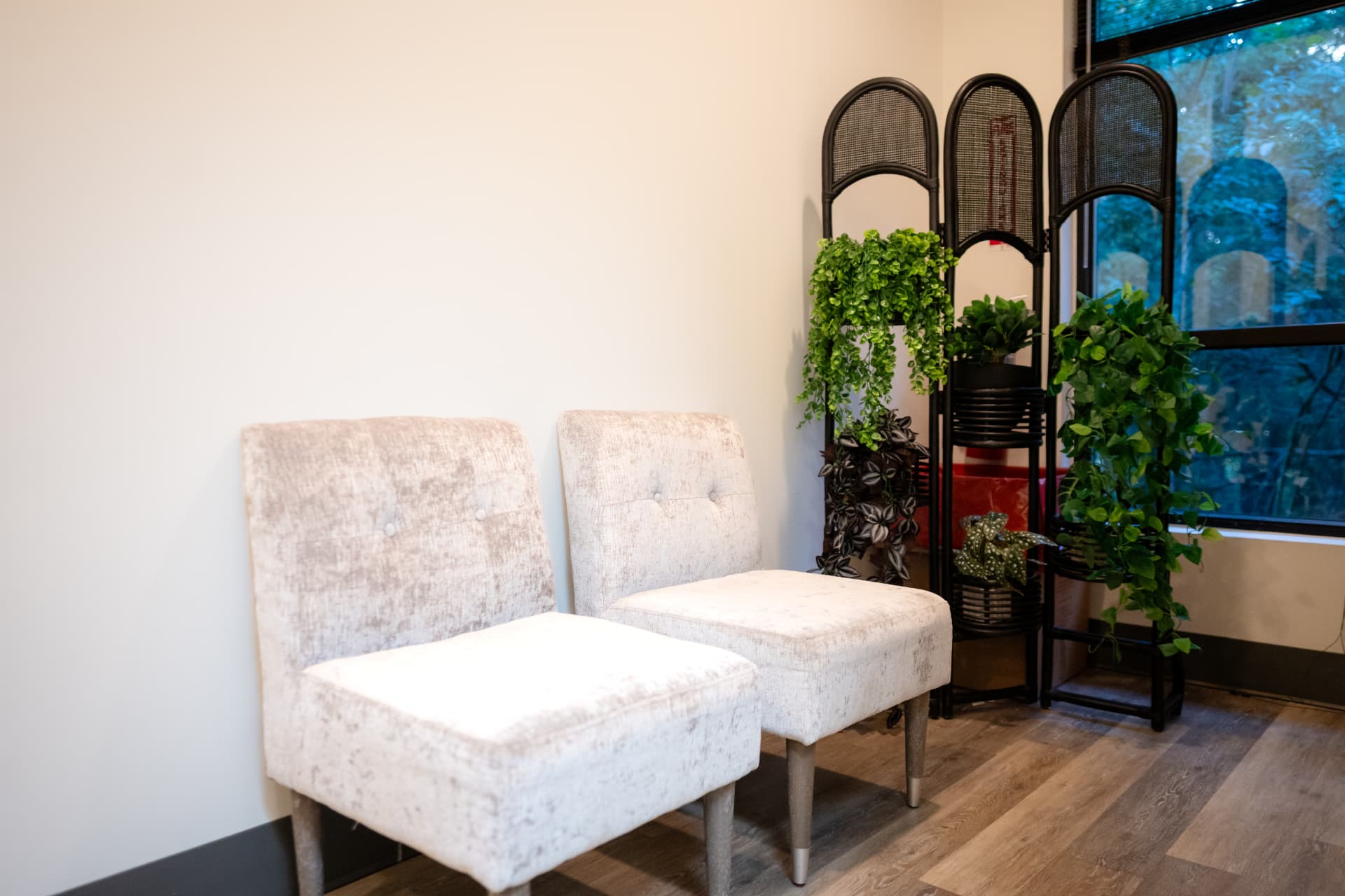 A quiet waiting area at Restora with two light upholstered chairs, plants, and a decorative screen near a window
